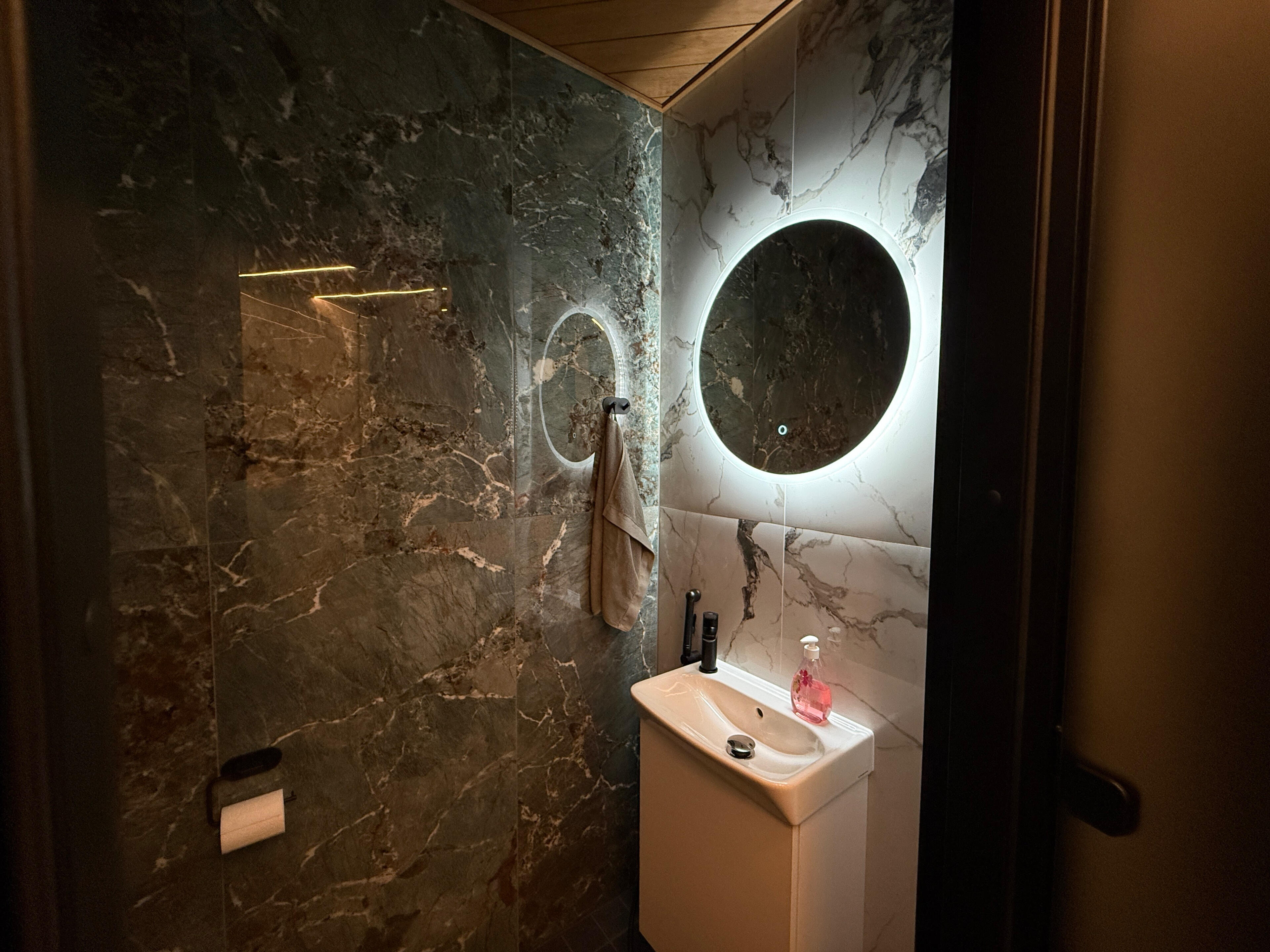 Vaaralan Saunan moderni wc-tila, jossa on marmorikuvioidut seinät, pesuallas ja valaistu pyöreä peili